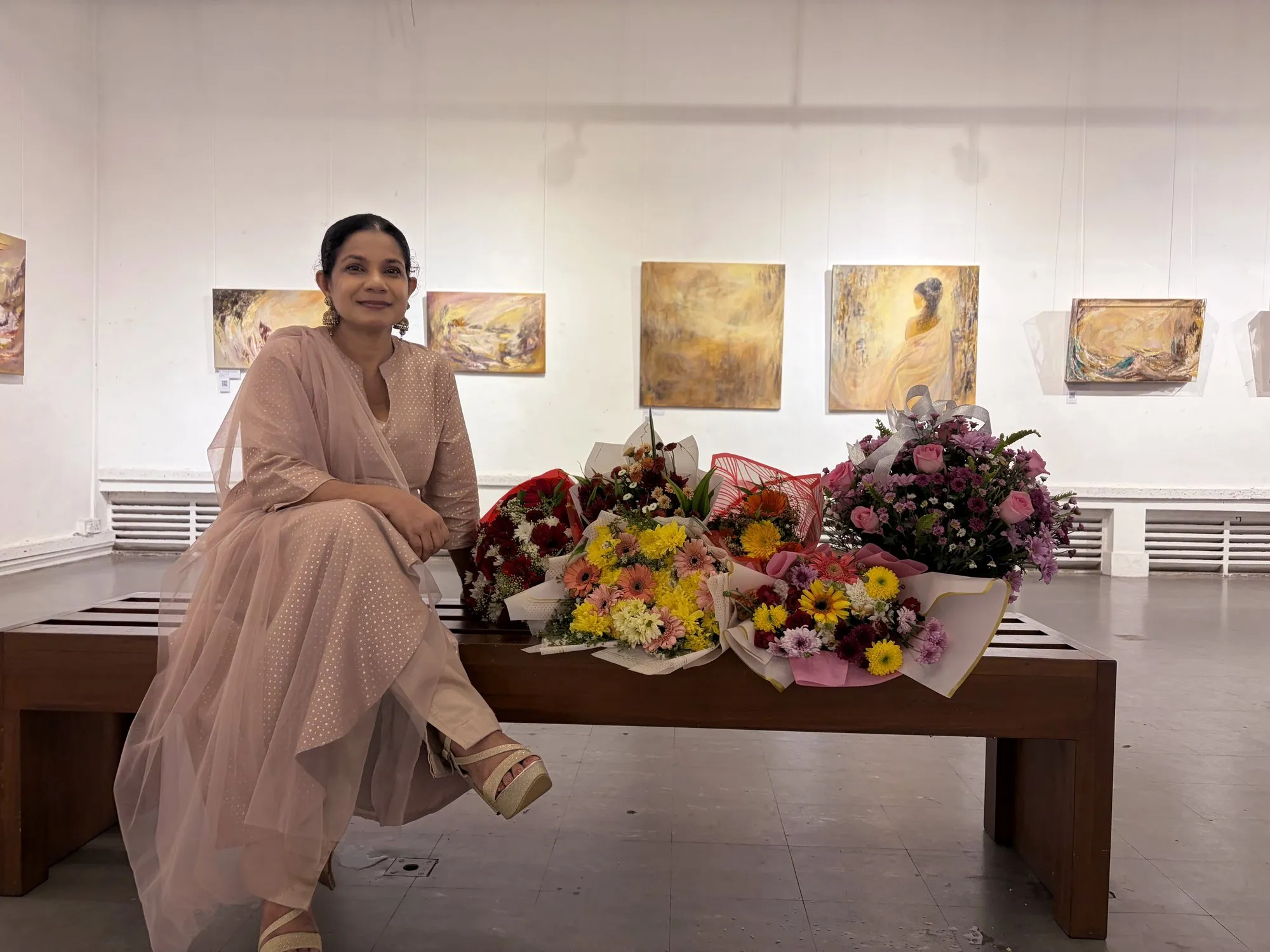 Priyadarshani Tennakoon seated among bouquets at her debut solo exhibition, her paintings on the gallery wall behind